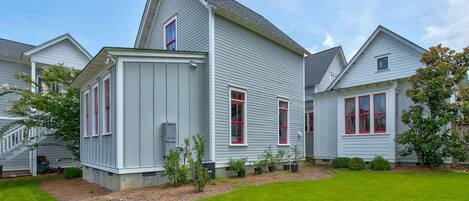 A two-story house with a sloped roof, white siding, and red-framed windows. There is a well-maintained lawn and a small garden area with various plants.