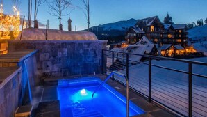 Un espace avec un bain à remous, doté d’un mur en pierre et d’une rambarde en métal, offrant une vue sur un paysage enneigé et sur un grand chalet composé de plusieurs bâtiments.