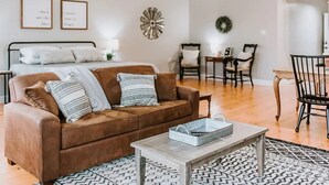 A living room with a brown sofa, a coffee table, a patterned rug, a dining area with chairs, and a bed with white bedding. - GrueneGusthaus#3| Private Condo+ Golf Cart Rentals (New Braunfels)