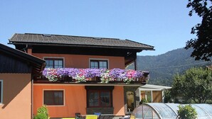 A two-story house with a balcony adorned with flowers, a greenhouse, and outdoor seating. - Comfortable Apartment Near Nassfeld (Hermagor-Pressegger See)