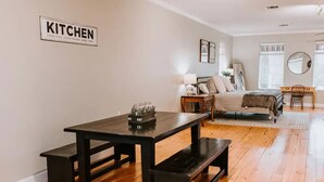Een moderne woonkamer met een donker houten eettafel, banken, een bank en een bed. Een bordje met de tekst 'Keuken' is aan de muur te zien.