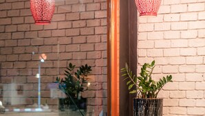 A bar area with a brick wall, two hanging red patterned lights, a potted plant, and white bar stools. - The Dorm Hostel (Phuket)