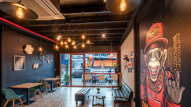 A modern cafe with wooden flooring, multiple seating options including tables and benches, and a large mural on the wall. - The Dorm Hostel (Phuket)