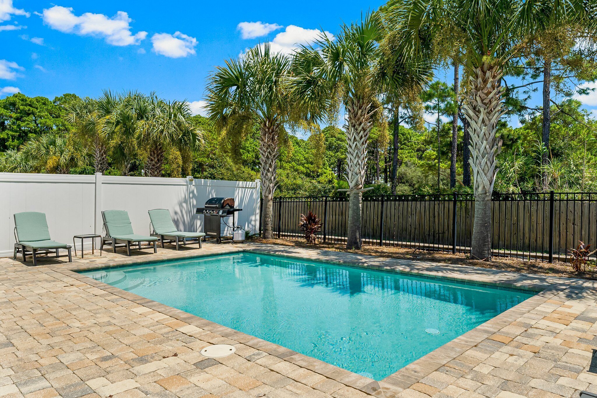 A rectangular swimming pool with a tiled edge, surrounded by a brick patio. There are three green lounge chairs aligned along the pool's edge.