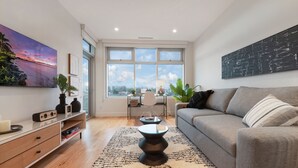 A modern living room with a grey sofa, a coffee table, a TV, and a large window with a city view. - West Bay Bliss (Victoria)