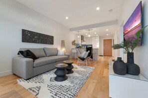 Una sala de estar moderna con un sofá gris, una mesa de centro y una alfombra. Se observa una cocina al fondo con un comedor.