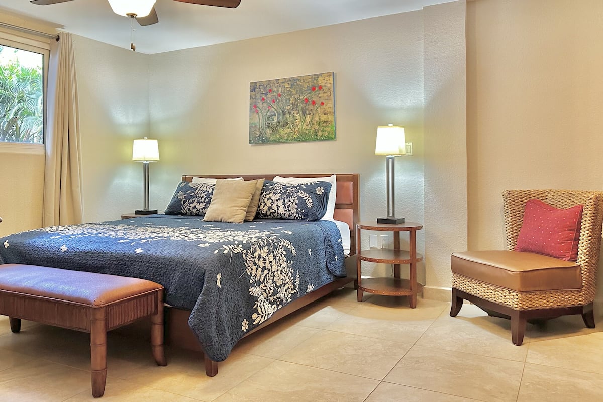 A bedroom with a bed, two bedside lamps, a ceiling fan, a bench, a chair, and a painting on the wall.