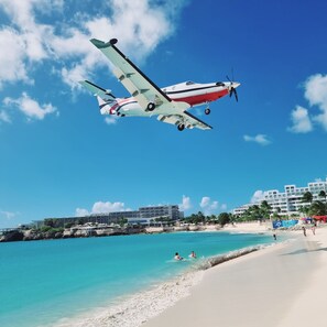 A beach with clear turquoise waters, a sandy shore, and a small propeller plane flying overhead. - OASIS Apartments Simpson Bay (Simpson Bay)