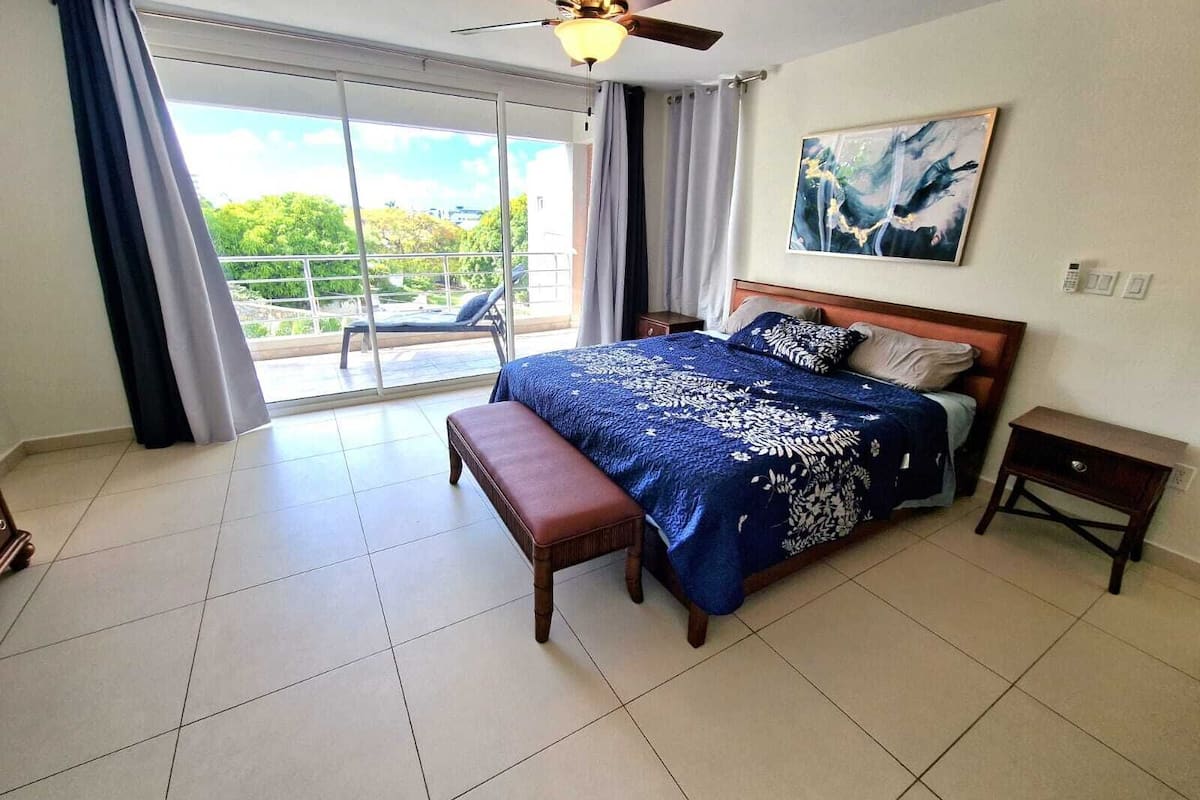 A bedroom with a bed, a bench, a bedside table, a ceiling fan, and a view of greenery through a sliding door.