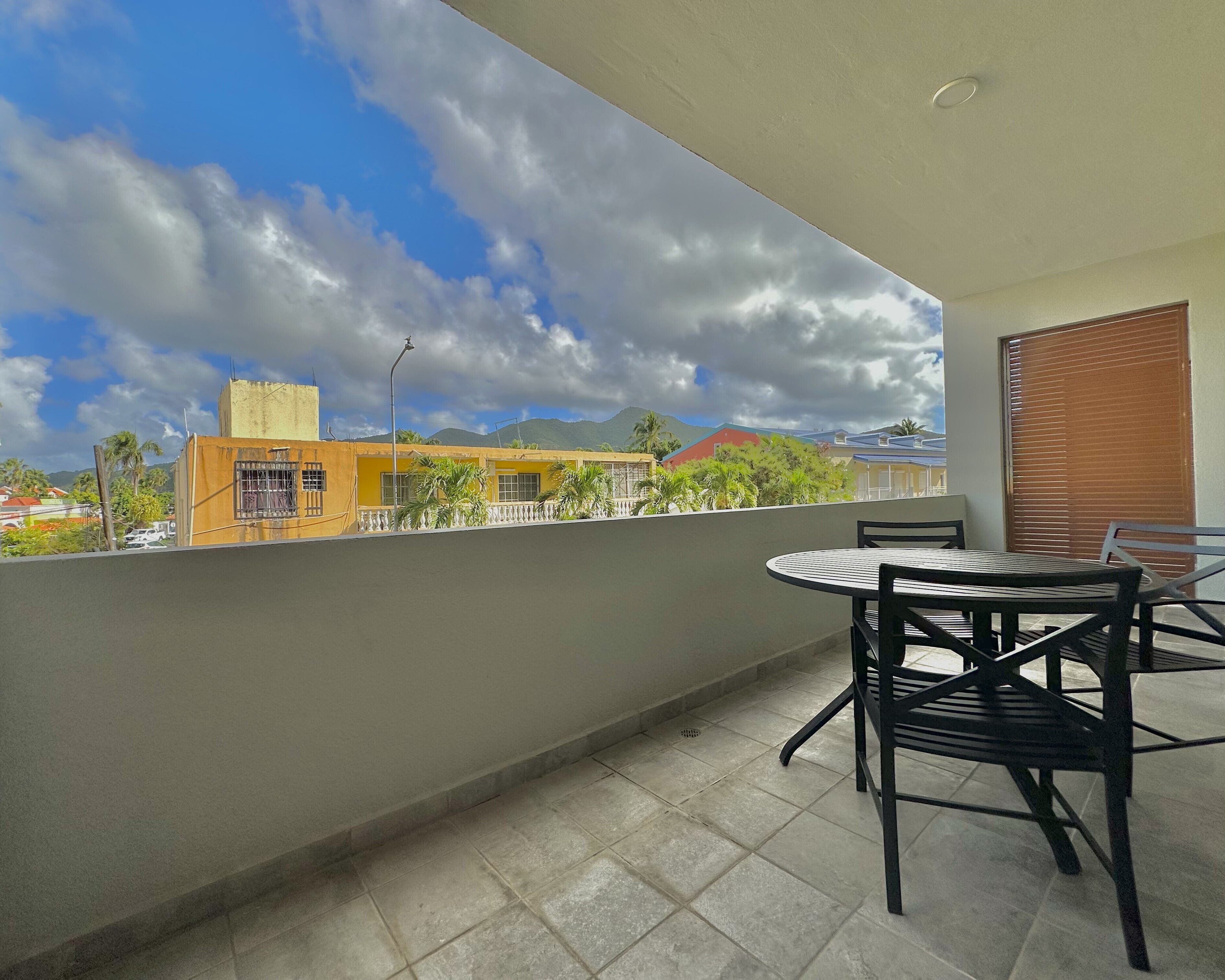 A balcony with a table and chairs, a view of buildings, and a cloudy sky.