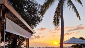Una terraza de madera con mobiliario exterior, una palmera y vistas al atardecer sobre el agua.