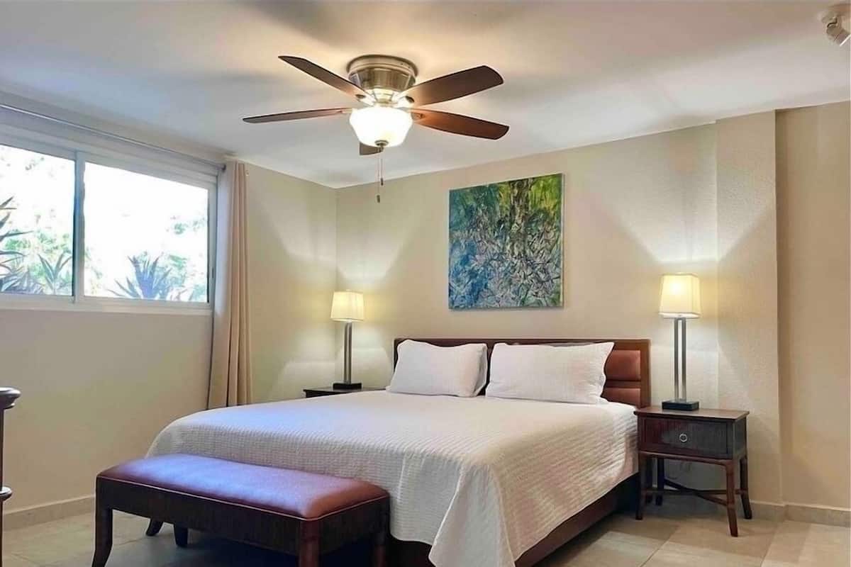 A neatly arranged bedroom with a bed, two bedside tables with lamps, a bench, a ceiling fan, and a window with curtains.