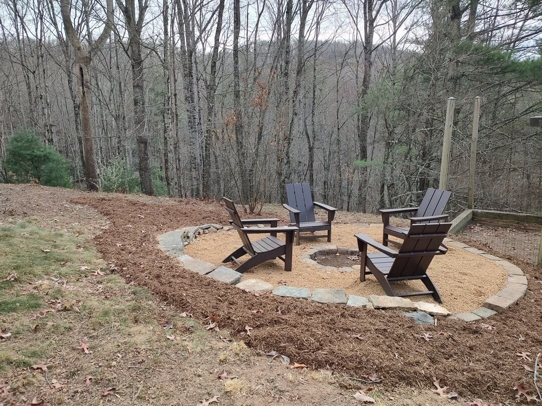 A circular seating area with wooden chairs and a fire pit, surrounded by a stone border, set in a wooded area.