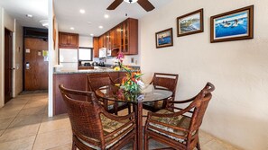 Un coin repas avec des chaises en osier et une table en verre, des meubles de cuisine en arrière-plan et des tableaux encadrés au mur.