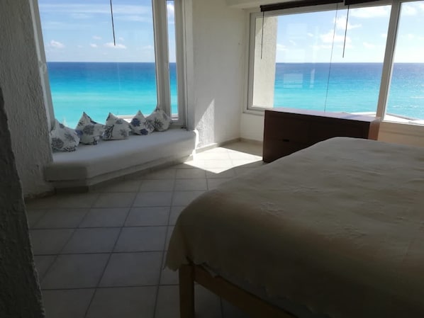 A bedroom with a large bed, a window with a view of the ocean, a bench with cushions, and a wooden nightstand.