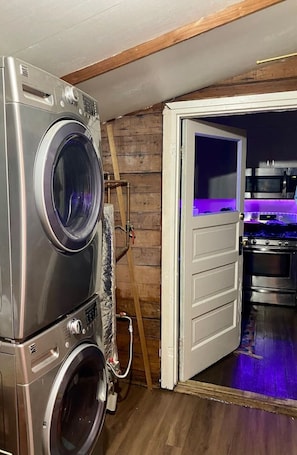 A laundry area with stacked washing machines, a wooden wall, and a kitchen visible through an open door.