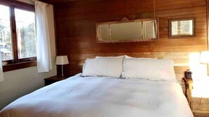 Quarto com teto de madeira, cama com roupa de cama branca, mesa de cabeceira com abajur e janela com cortinas.