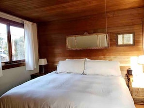 A bedroom with a wooden ceiling, a bed with white bedding, a nightstand with a lamp, and a window with curtains. - Beat the Heat in our Sunny Classic Inverness Home (Inverness)