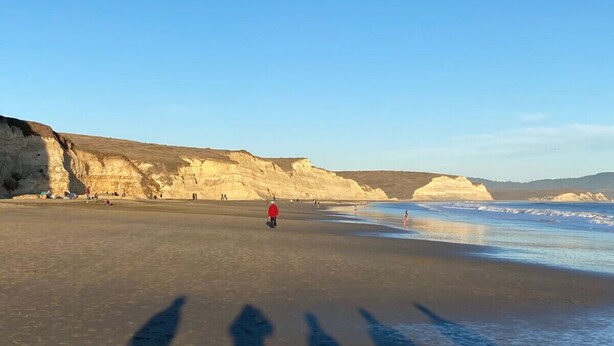 Uma praia com uma pessoa num casaco vermelho caminhando em direção à água, falésias ao fundo e sombras de pessoas na areia.