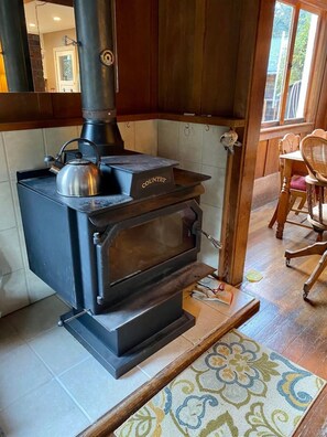 A wood-burning stove with a kettle on top, a wooden table with chairs, and a patterned rug on the floor.