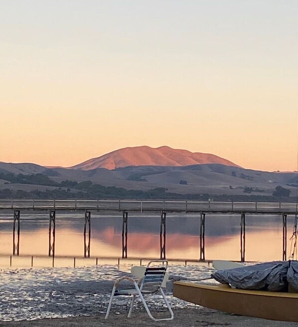 A serene lakeside view with a mountain in the background, a dock with a boat, and a lounge chair.