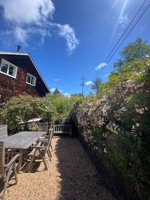 A wooden deck with a table and chairs, surrounded by dense foliage and a brick house.
