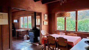 Interior rústico em madeira, com área de jantar, lareira e grandes janelas que dão para o verde.