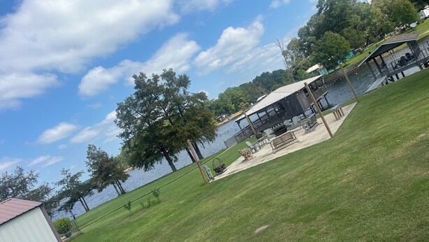 A lakeside property with a grassy lawn, a covered dock, and a small building. - Lake front home in quiet neighborhood & mins away from boat launch and amenities (Many)