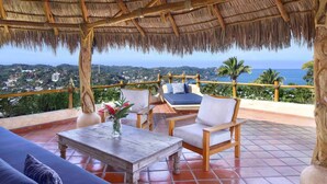A terrace with a thatched roof, wooden furniture, and a view of the ocean.