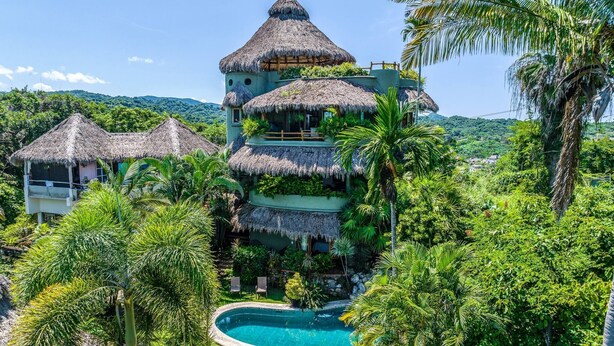 A multi-story resort with thatched-roof structures, a swimming pool, and lush tropical vegetation.