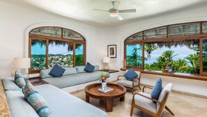 A spacious living room with a large sofa, armchairs, and a coffee table. There are two large windows offering a view of the sea and greenery.