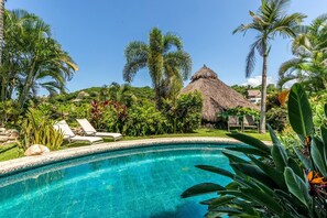 Una piscina azul cristalina rodeada de exuberante vegetación y un edificio con techo de paja.