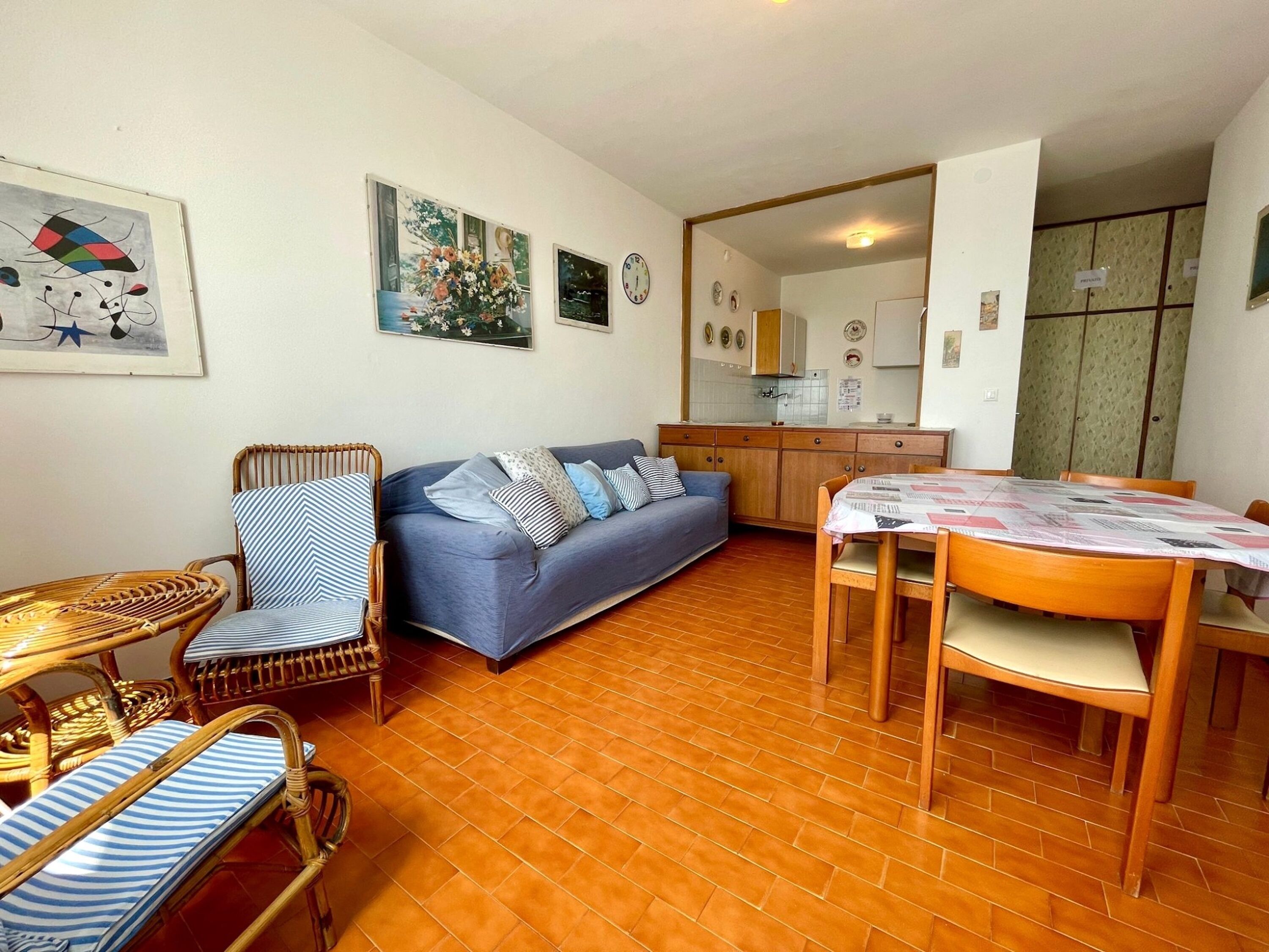 A cozy living room with a sofa, wicker chairs, a dining table, and a kitchen in the background.