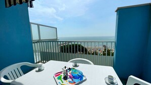 Ein Balkon mit einem weißen Tisch, Stühlen und einer Ablage mit Kaffeekanne und Tassen. Durch das Geländer ist Meer und Strand zu sehen.