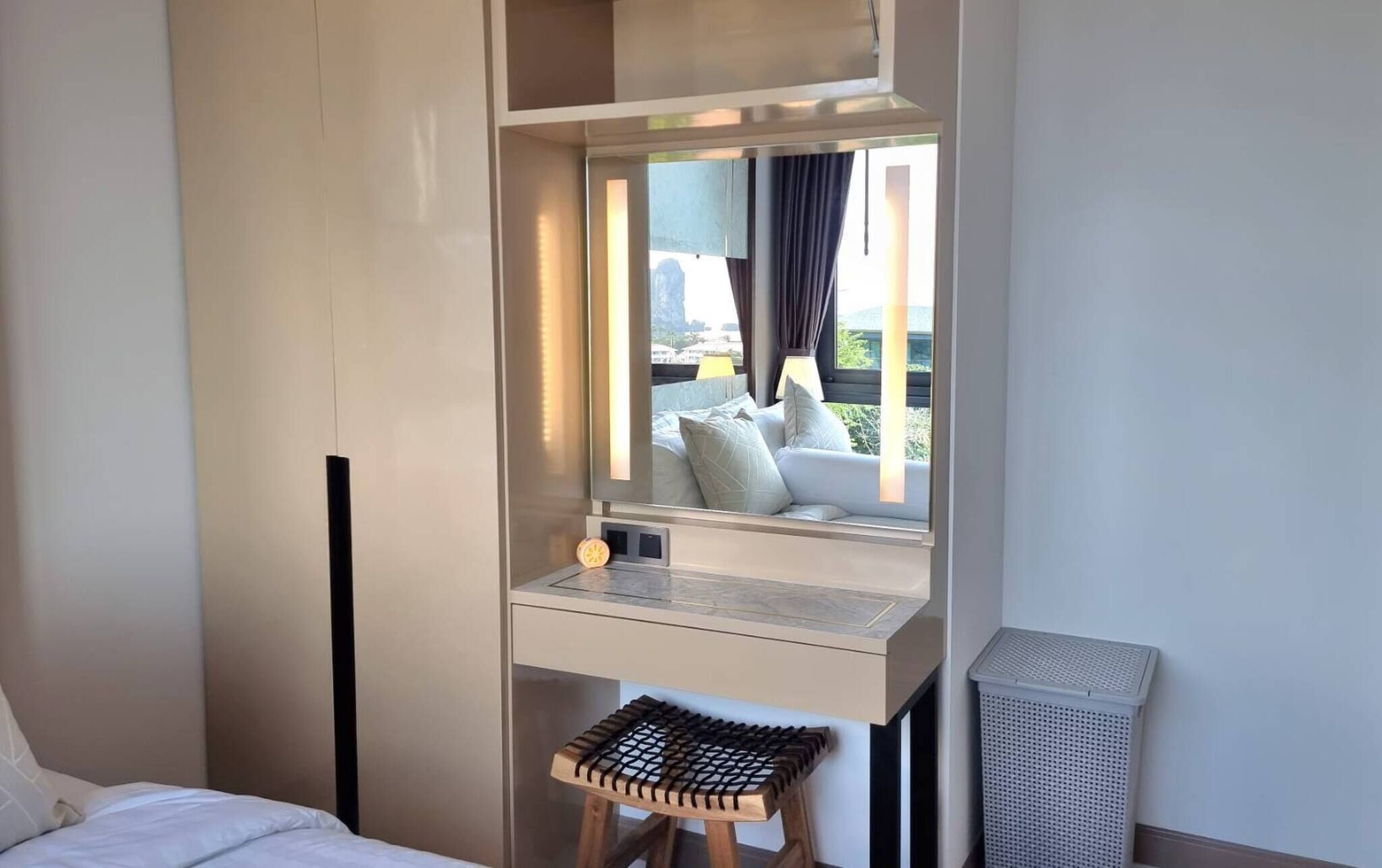 Ein Hotelzimmer mit Bett, einem kleinen Hocker, einem Schreibtisch mit Lampe und einem Fenster mit Blick auf Gebäude und Grünflächen.