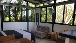 A room with a bed, a sofa, a dining table, and a chair. There are large windows with a view of trees outside.