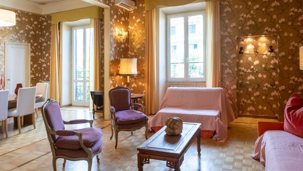 A room with a checkered floor, a wooden coffee table, purple armchairs, a pink sofa, and a ceiling with decorative panels.