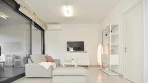 A modern living room with a sofa, ottoman, TV, white shelves, and a glass door leading to a balcony.