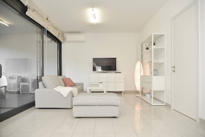 A modern living room with a sofa, ottoman, TV, and white shelving unit.