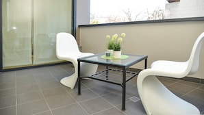 A modern outdoor seating area with a glass table and two white chairs, a small potted plant, and a view of a building and trees.
