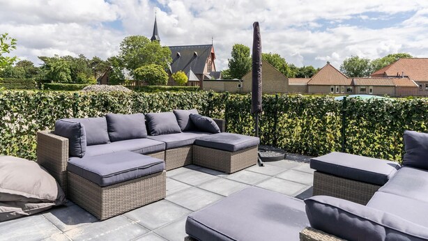 An outdoor seating area with wicker furniture and cushions, a hedge, and a building with a church in the background. - Group Apartment on Ameland by De Vleijen (Nes)