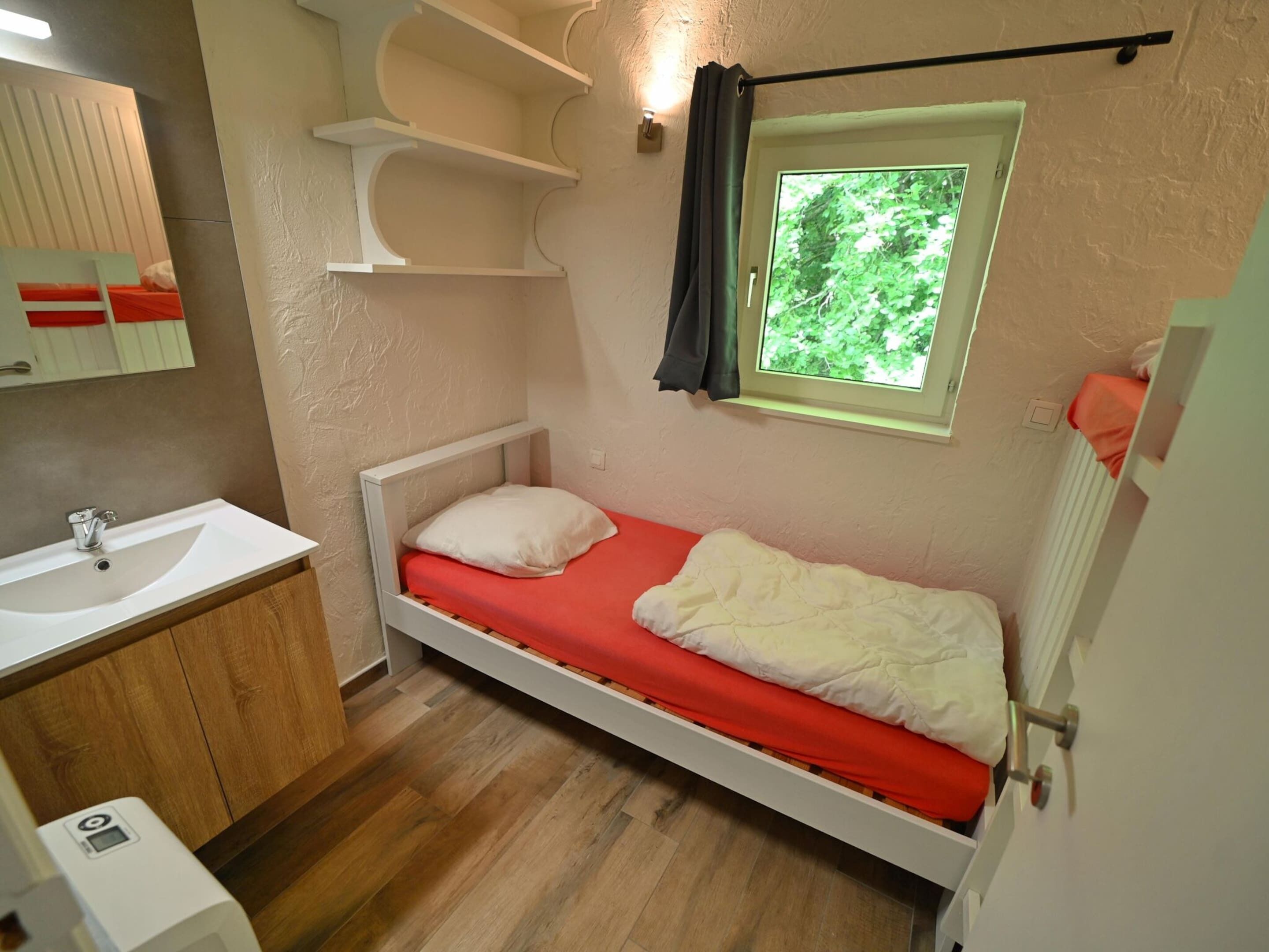 A small, single-bed room with a red mattress, a white pillow, a wooden cabinet with a sink, and a window with a view of greenery.
