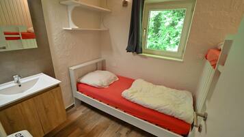 A small, single-bed room with a red mattress, a white pillow, a wooden cabinet with a sink, and a window with a view of greenery.