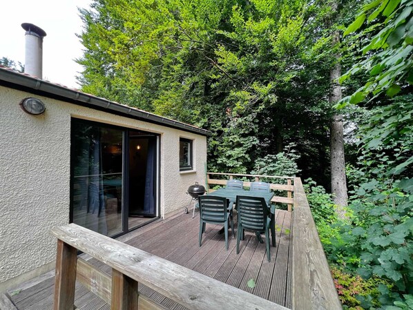 A wooden deck with a table and chairs, a small building with a chimney, and a glass door leading to an interior space. - Bungalow in Virton by Vallee de Rabais Lake (Virton)