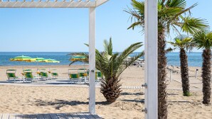 A beach with a wooden pergola, palm trees, and beach chairs under umbrellas. - Cozy apartment in Cavallino-Treporti (Cavallino-Treporti)