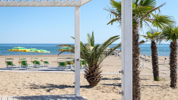 A beach with a wooden pergola, palm trees, and beach chairs under umbrellas. - Cozy apartment in Cavallino-Treporti (Cavallino-Treporti)