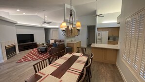 Un salon moderne avec une cheminée, un coin repas avec une longue table et une cuisine équipée d’un réfrigérateur et de placards.