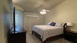 Une chambre à coucher avec un lit, un ventilateur de plafond, un téléviseur posé sur un meuble et une fenêtre avec des rideaux.
