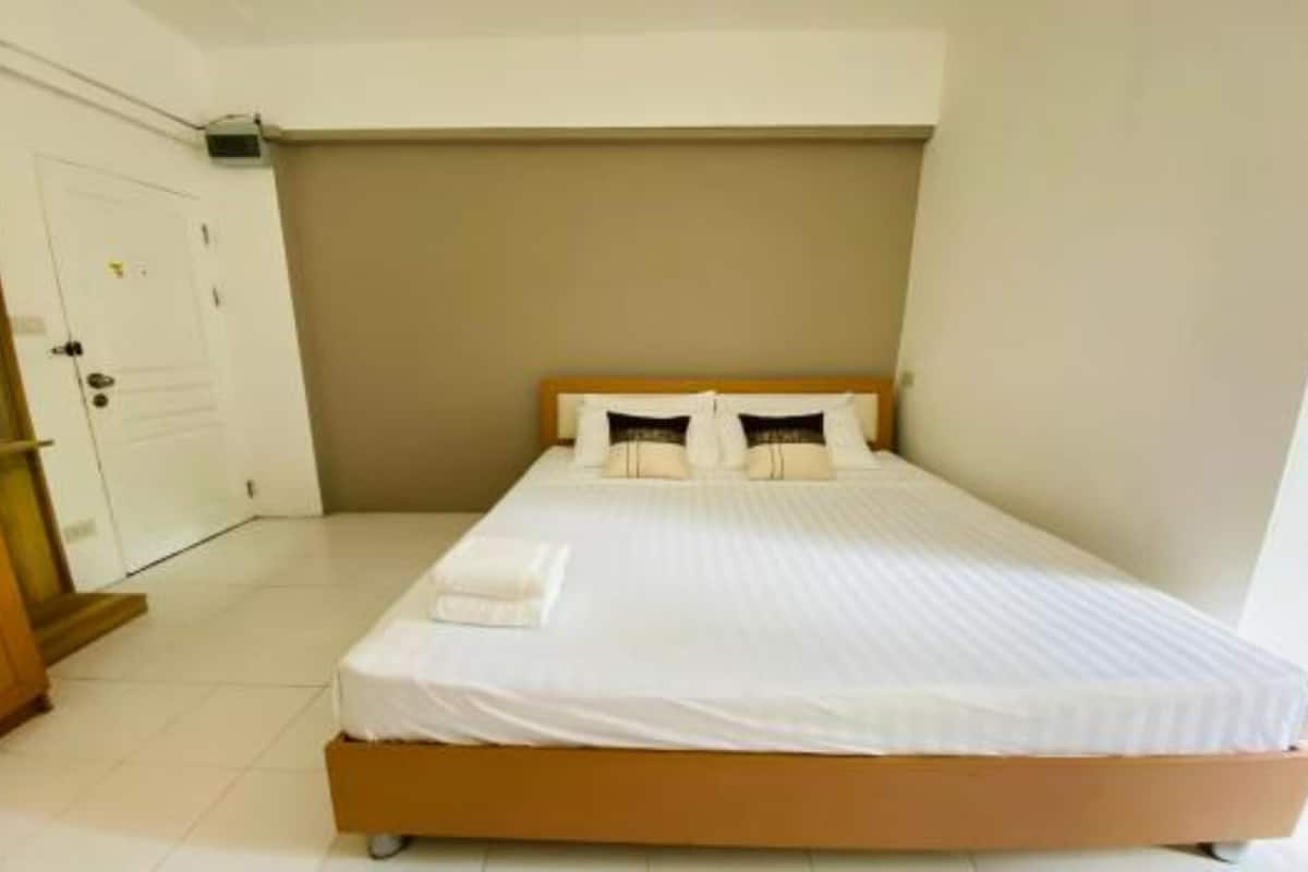 Un lit simple avec une literie blanche et deux oreillers, une tête de lit en bois et une porte blanche, dans une pièce aux murs blancs et au sol carrelé.