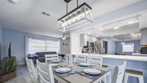 A modern kitchen with white cabinets, a central island, and a dining area with a wooden table and white chairs. - 🏝️10 MINS TO BEACH # HEATED POOL/SPA # SLEEP FOR 12@ KIDS FRIENDLY (Melbourne)